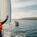 En person iklädd flytväst på en segelbåt vinkar till en mötande båt.